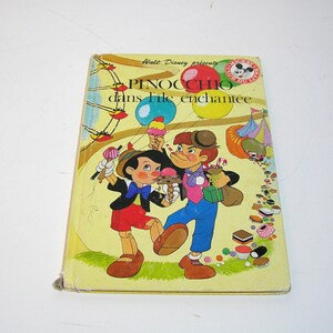 May include: Vintage children's book titled "Pinocchio dans l'île enchantée" featuring illustrations of Pinocchio and other characters. The cover has a yellow background with colorful images of ice cream, balloons, and a circus tent. The book is a Walt Disney production.