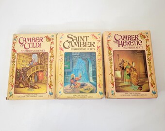 May include: Three vintage paperback books with fantasy art covers. Titles include "Camber of Culdi," "Saint Camber," and "Camber the Heretic" by Katherine Kurtz. The covers feature illustrations of medieval scenes with figures and architectural elements.