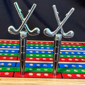 Könnte beinhalten: Zwei silberne, golfclubförmige Stecknadeln sind in ein hölzernes Spielbrett mit roten, grünen und blauen Quadraten gesteckt. Das Brett hat ein Gitter aus Löchern.