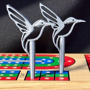 Op de afbeelding: Twee zilveren spelstukken in de vorm van een kolibrie. De kolibries zitten op cilindrische palen. De spelstukken staan op een houten bord met een kleurrijke baan en het woord "START".