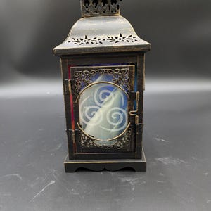 May include: A decorative black metal lantern with a distressed gold finish. The lantern features a glass panel with a white spiral design and ornate metalwork. The top has a small handle and decorative cutouts.