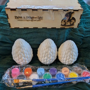 May include: A set of three white ceramic dragon eggs with a scaly texture, paintbrushes, and a set of 10 paint pots in various colors. The paint pots are labeled with the colors: pink, orange, light blue, purple, brown, yellow, and red. The box in the background reads "Paint a Dragon Egg" and "contains 5 dragon eggs and paint".