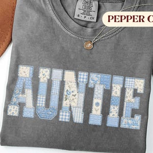 May include: A pepper-colored short-sleeve t-shirt featuring the word AUNTIE in a blue and white patchwork-style floral and gingham print. The shirt has a Comfort Colors brand label at the neck and a small gold-toned circular pendant necklace.