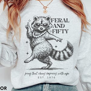 May include: Ash-colored sweatshirt featuring a raccoon wearing sunglasses and a pearl necklace. The graphic includes the text "FERAL AND FIFTY" and "proof that chaos improves with age EST. 1976". The sweatshirt is a light gray color.