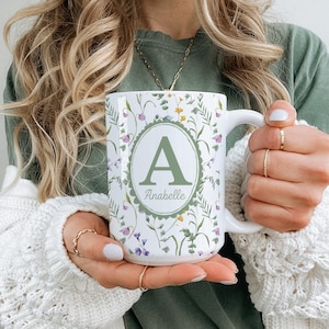 May include: White ceramic mug with a floral design and the letter "A" in a green oval. The mug features the name "Anabelle" below the letter. The floral pattern includes purple, yellow, and green accents.
