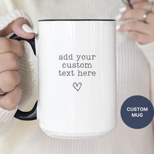 May include: White ceramic mug with a black handle and rim. The mug features the text "add your custom text here" and a heart symbol. A navy blue circle with the words "CUSTOM MUG" is also visible.