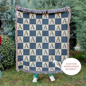 May include: A custom monogram blanket with a checkerboard pattern in navy blue and tan, featuring the letter "A". The blanket has a fringed edge and the text "CUSTOM MONOGRAM BLANKET".