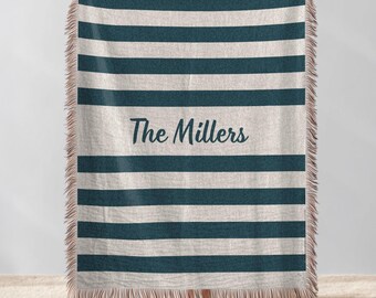 May include: A beige and teal striped throw blanket with fringed edges. The blanket has horizontal teal stripes and the words "The Millers" in a script font. The blanket is held up by a person.
