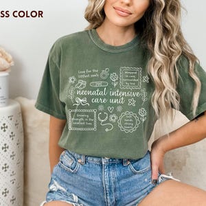Op de afbeelding: Mosgroen t-shirt met witte tekst en illustraties over neonatale intensive care. De tekst bevat zinnen als "Love for the littlest souls" en "neonatal intensive care unit". Het shirt wordt door een persoon gedragen.
