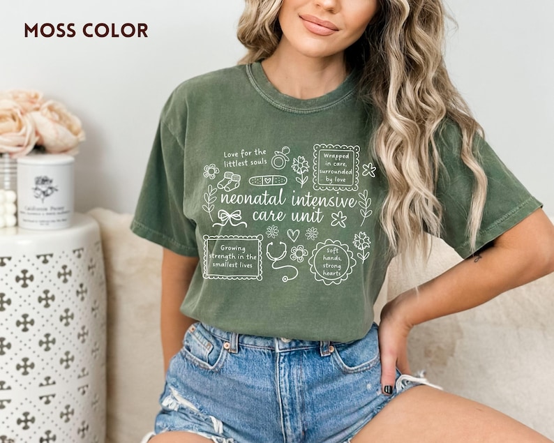 Op de afbeelding: Mosgroen t-shirt met witte tekst en illustraties over neonatale intensive care. De tekst bevat zinnen als "Love for the littlest souls" en "neonatal intensive care unit". Het shirt wordt door een persoon gedragen.