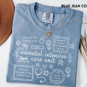 Op de afbeelding: Een blauw spijkerbroekkleurig T-shirt met witte tekst en illustraties over neonatale intensive care. Het shirt bevat zinnen als "Love for the littlest souls" en "neonatal intensive care unit". Een gouden ketting ligt over het shirt.