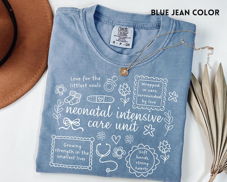 Op de afbeelding: Een blauw spijkerbroekkleurig T-shirt met witte tekst en illustraties over neonatale intensive care. Het shirt bevat zinnen als "Love for the littlest souls" en "neonatal intensive care unit". Een gouden ketting ligt over het shirt.