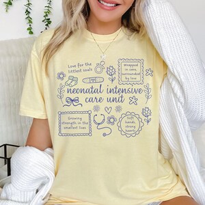 Op de afbeelding: Een lichtgeel t-shirt met de woorden "neonatal intensive care unit" in blauw schrift. Het shirt heeft blauwe illustraties van een stethoscoop, bloemen en de tekst "Love for the littlest souls" en "Growing strength in the smallest lives."