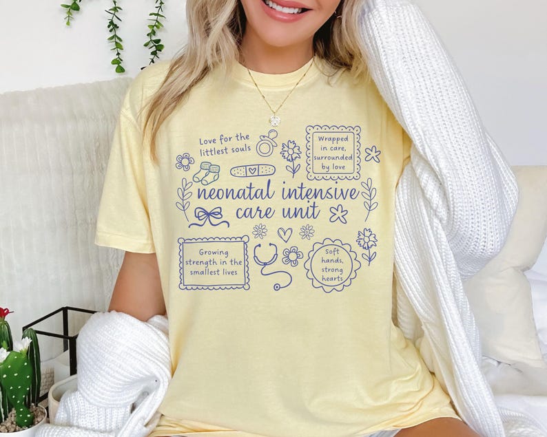 Op de afbeelding: Een lichtgeel t-shirt met de woorden "neonatal intensive care unit" in blauw schrift. Het shirt heeft blauwe illustraties van een stethoscoop, bloemen en de tekst "Love for the littlest souls" en "Growing strength in the smallest lives."