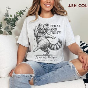 Puede incluir: Camiseta color ceniza con un gr&aacute;fico de un mapache con gafas de sol y un collar de perlas, con el texto "FERAL AND FORTY". Debajo del mapache est&aacute; el texto "Is my 40th Birthday AUSTIN, TX 2026".