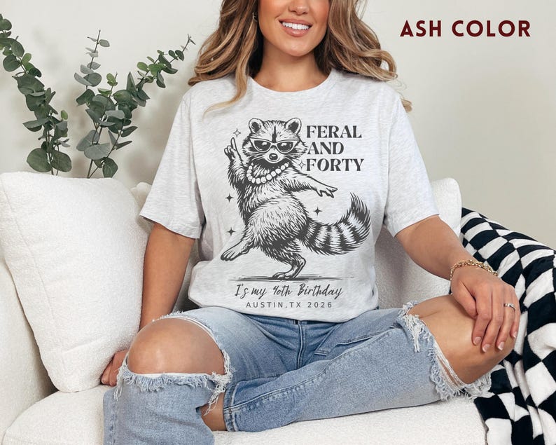 Puede incluir: Camiseta color ceniza con un gr&aacute;fico de un mapache con gafas de sol y un collar de perlas, con el texto "FERAL AND FORTY". Debajo del mapache est&aacute; el texto "Is my 40th Birthday AUSTIN, TX 2026".