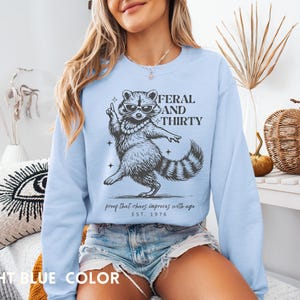 May include: Light blue sweatshirt with a raccoon graphic wearing sunglasses and a pearl necklace. The text reads "FERAL AND THIRTY" with the phrase "proof that chaos improves with age" and "EST. 1976".