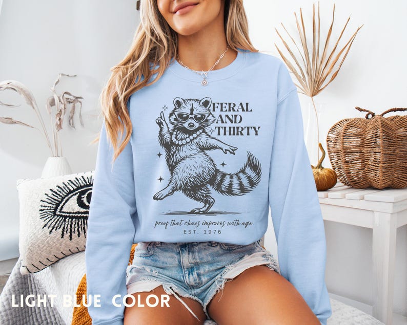 May include: Light blue sweatshirt with a raccoon graphic wearing sunglasses and a pearl necklace. The text reads "FERAL AND THIRTY" with the phrase "proof that chaos improves with age" and "EST. 1976".