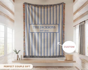 May include: A woven throw blanket with blue vertical stripes and a rectangular central panel. The panel displays the text "THE JACKSONS EST 2025". The blanket has a fringed edge and is displayed on a wall.
