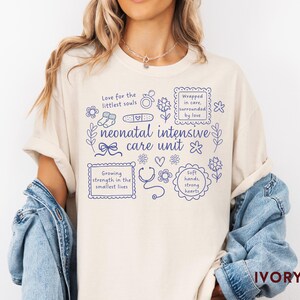 Op de afbeelding: Ivoor kleurig T-shirt met blauwe tekst en illustraties over een neonatale intensive care unit. Het ontwerp bevat zinnen als "Love for the littlest souls" en "Growing strength in the smallest lives."