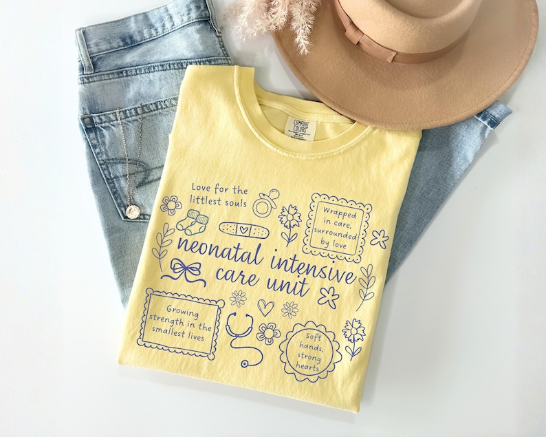 Op de afbeelding: Een geel t-shirt met blauwe tekst en illustraties, waaronder de woorden "neonatal intensive care unit". Het shirt ligt op een blauwe spijkerbroek en naast een beige hoed. Het shirt heeft verschillende zinnen en illustraties.