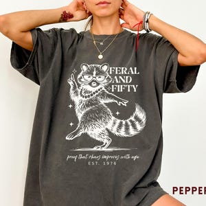 May include: Dark grey t-shirt with a white graphic of a raccoon wearing glasses and a pearl necklace, with the text "FERAL AND FIFTY" above the raccoon. Below the raccoon is the text "proof that chaos improves with age EST. 1976".