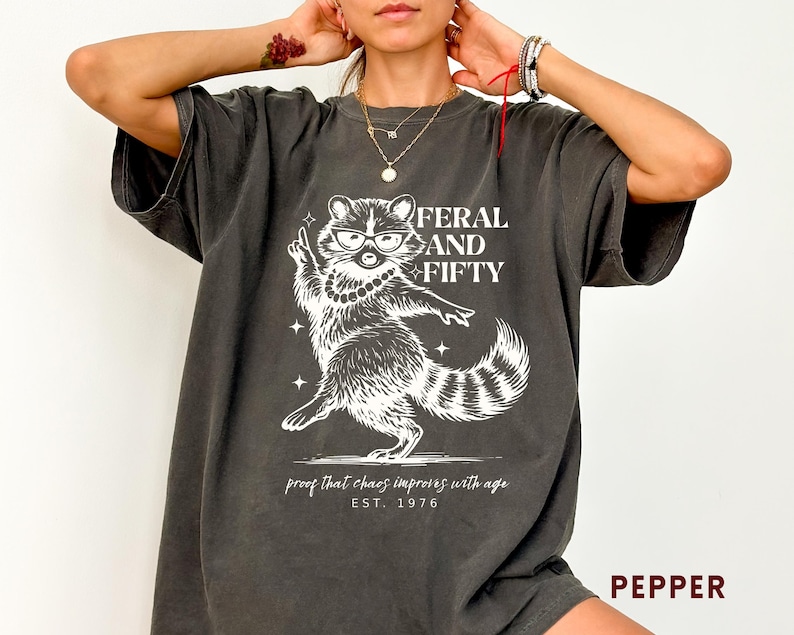 May include: Dark grey t-shirt with a white graphic of a raccoon wearing glasses and a pearl necklace, with the text "FERAL AND FIFTY" above the raccoon. Below the raccoon is the text "proof that chaos improves with age EST. 1976".