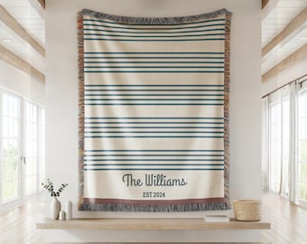 May include: A woven throw blanket featuring a cream-colored base with horizontal dark blue stripes. The bottom section displays the text "The Williams" in a script font above the text "EST 2024" in a sans-serif font, with fringed edges.