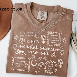 Op de afbeelding: Bruin T-shirt met witte tekst en illustraties, waaronder de woorden "neonatal intensive care unit". Het ontwerp van het shirt bevat zinnen als "Wrapped in care, surrounded by love".