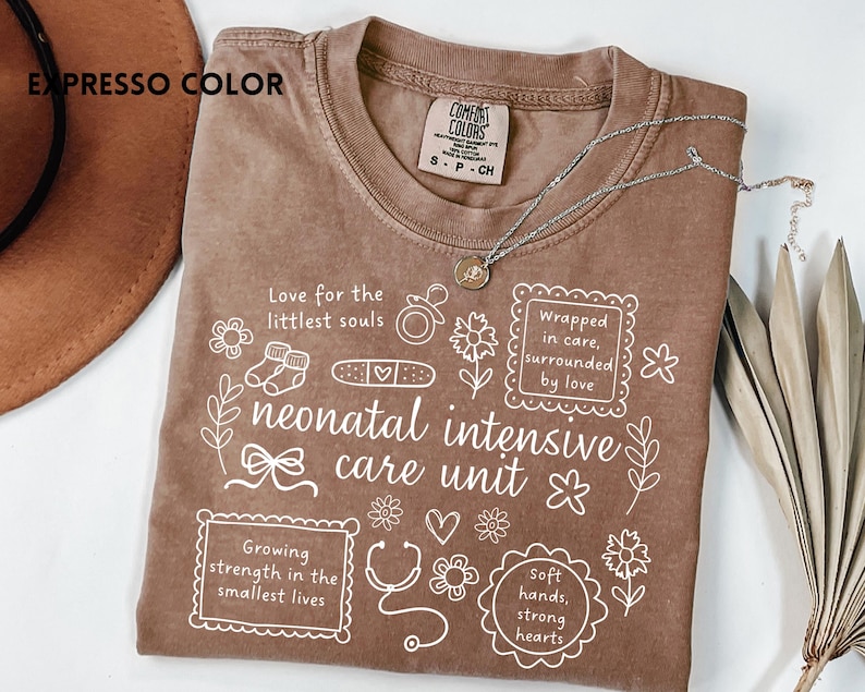 Op de afbeelding: Bruin T-shirt met witte tekst en illustraties, waaronder de woorden "neonatal intensive care unit". Het ontwerp van het shirt bevat zinnen als "Wrapped in care, surrounded by love".