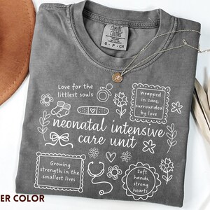 Op de afbeelding: Pepperkleurig t-shirt met witte grafische tekst en illustraties over een neonatale intensive care unit. Het shirt bevat zinnen als "Liefde voor de kleinste zielen" en "Omringd door zorg, omringd door liefde".