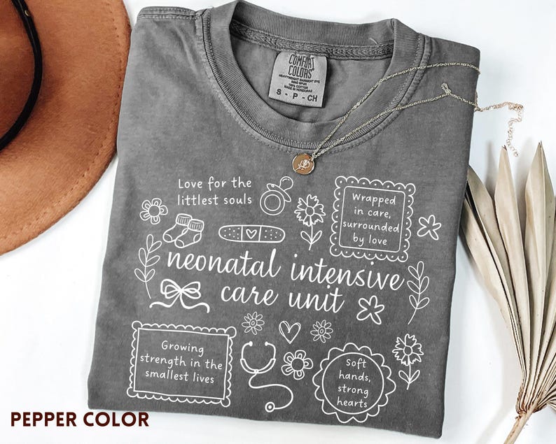 Op de afbeelding: Pepperkleurig t-shirt met witte grafische tekst en illustraties over een neonatale intensive care unit. Het shirt bevat zinnen als "Liefde voor de kleinste zielen" en "Omringd door zorg, omringd door liefde".