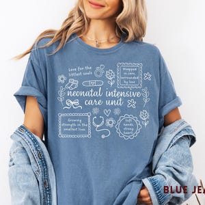 Op de afbeelding: Blauw spijkerbroekkleurig T-shirt met witte tekst en illustraties over neonatale intensive care. Het shirt bevat zinnen als "Love for the littlest souls" en "Growing strength in the smallest lives."