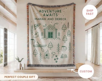 May include: A beige woven throw blanket with a green camping-themed design. The design includes a cabin, trees, a boot, a canoe, and the words "ADVENTURE AWAITS" and a personalized name. The blanket has a fringed edge.