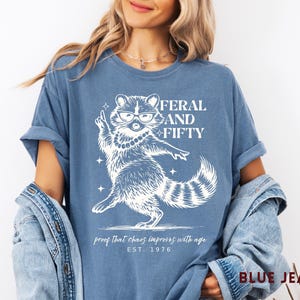May include: Blue jean-colored t-shirt with a white graphic of a raccoon wearing glasses and a pearl necklace. The text reads "FERAL AND FIFTY" above the raccoon, with "proof that chaos improves with age" and "EST. 1976" below.