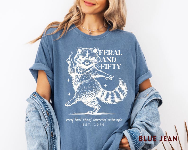 May include: Blue jean-colored t-shirt with a white graphic of a raccoon wearing glasses and a pearl necklace. The text reads "FERAL AND FIFTY" above the raccoon, with "proof that chaos improves with age" and "EST. 1976" below.