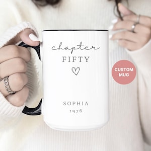 May include: White ceramic mug with black handle and rim, featuring the words "chapter FIFTY" with a heart symbol, the name "SOPHIA", and the year "1976". A pink circle with the words "CUSTOM MUG" is also visible.