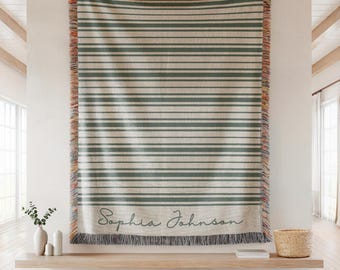May include: A woven throw blanket with horizontal green stripes on a cream background. The blanket has a fringed edge with orange and gray accents. The name "Sophia Johnson" is written in cursive at the bottom of the blanket.