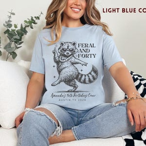 Puede incluir: Camiseta azul claro con un gr&aacute;fico de un mapache con gafas de sol y un collar de perlas, con el texto "FERAL AND FORTY". Debajo del mapache est&aacute; el texto "Amanda's 40th Birthday Crew AUSTIN, TX 2026".
