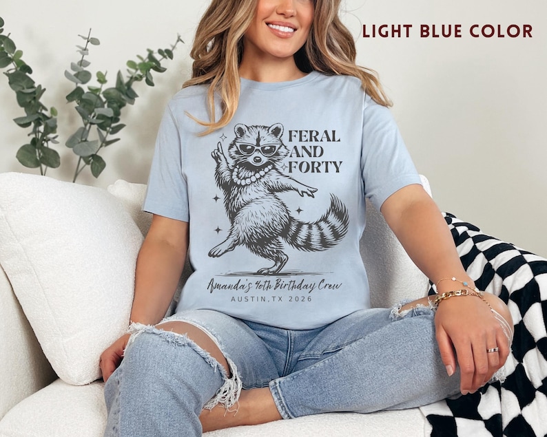 Puede incluir: Camiseta azul claro con un gr&aacute;fico de un mapache con gafas de sol y un collar de perlas, con el texto "FERAL AND FORTY". Debajo del mapache est&aacute; el texto "Amanda's 40th Birthday Crew AUSTIN, TX 2026".
