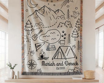 May include: A beige throw blanket with a black camping-themed design. The design includes mountains, trees, a tent, and the text "Mariah and Dereck Est 2025". The blanket has a fringed edge with orange, gray, and beige stripes.
