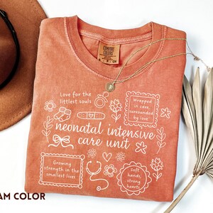 Op de afbeelding: Een koraalkleurig T-shirt met witte tekst en afbeeldingen gerelateerd aan neonatale intensive care. Het shirt bevat zinnen als "Love for the littlest souls" en "neonatal intensive care unit". Een gouden ketting en een bruine hoed zijn ook zichtbaar.