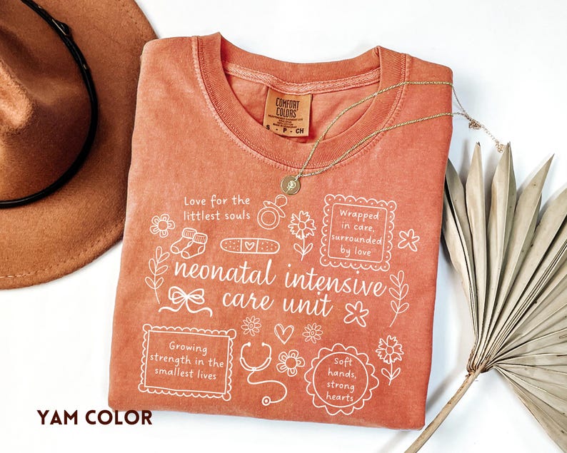 Op de afbeelding: Een koraalkleurig T-shirt met witte tekst en afbeeldingen gerelateerd aan neonatale intensive care. Het shirt bevat zinnen als "Love for the littlest souls" en "neonatal intensive care unit". Een gouden ketting en een bruine hoed zijn ook zichtbaar.