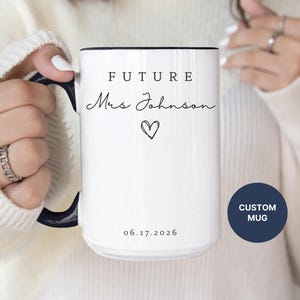 May include: White ceramic mug with black handle and rim. The mug features the text "FUTURE Mrs Johnson" with a heart symbol and the date "06.17.2026". A navy blue circle with the text "CUSTOM MUG" is also visible.
