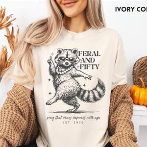 May include: Ivory-colored t-shirt featuring a raccoon wearing sunglasses and a pearl necklace. The text reads "FERAL AND FIFTY" with the phrase "proof that chaos improves with age" and "EST. 1976". The shirt is part of a clothing collection.