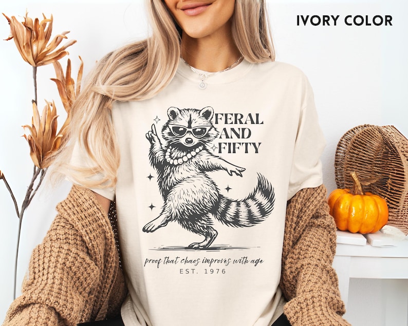 May include: Ivory-colored t-shirt featuring a raccoon wearing sunglasses and a pearl necklace. The text reads "FERAL AND FIFTY" with the phrase "proof that chaos improves with age" and "EST. 1976". The shirt is part of a clothing collection.