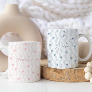 May include: Two white ceramic mugs with curved handles. One mug is light pink with a pattern of pink hearts and the name "Amelia" in cursive. The other mug is light blue with a pattern of blue hearts and the name "Amelia" in cursive.