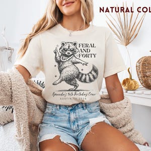 Puede incluir: Camiseta color crema con un gr&aacute;fico negro de un mapache con gafas de sol y collar de perlas, con el texto "FERAL AND FORTY". Debajo del mapache est&aacute; el texto "Amanda's 40th Birthday Crew, Austin, TX 2026".