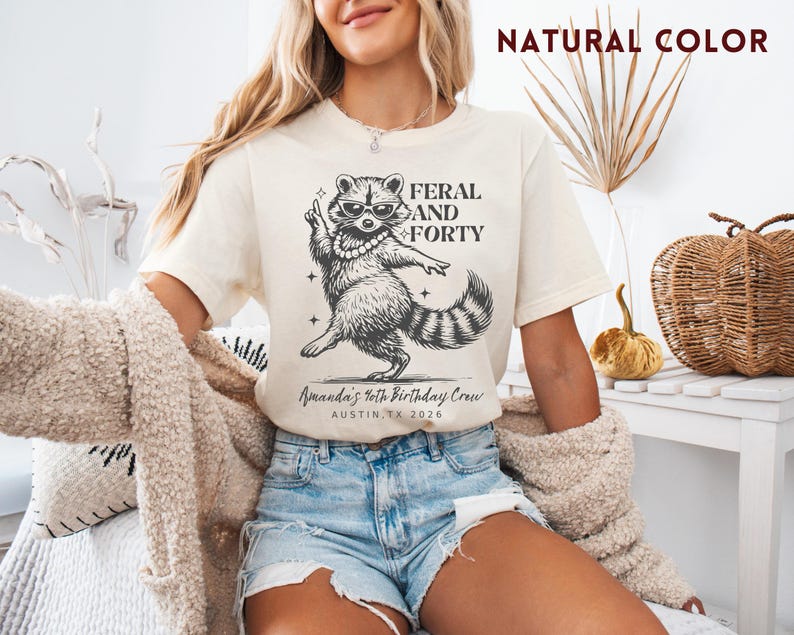 Puede incluir: Camiseta color crema con un gr&aacute;fico negro de un mapache con gafas de sol y collar de perlas, con el texto "FERAL AND FORTY". Debajo del mapache est&aacute; el texto "Amanda's 40th Birthday Crew, Austin, TX 2026".