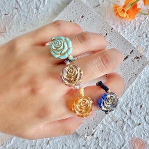 Puede incluir: Cuatro anillos con colgantes en forma de rosa en varios colores: turquesa, dorado, ámbar y azul. Los anillos están en una mano, mostrando los diferentes diseños y colores. Los anillos están hechos de cuentas y metal.